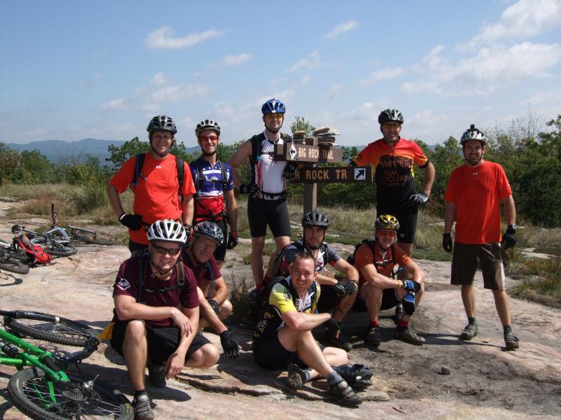 A group of eight mountain bikers posing together at a trail sign that points to "Big Rock Trail" and "Rock Trail." They are dressed in cycling gear and helmets, with several bicycles in the background. The setting is an outdoor area with grassy terrain and mountains in the distance under a partly cloudy sky. DuPont State Recreational Forest mountain bike trail.
