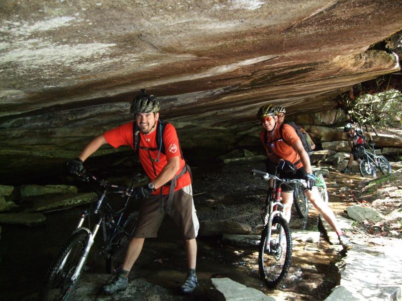 Two mountain bikers in bright shirts are maneuvering their bikes under a rocky overhang in a forested area. One biker is smiling while pushing their bike, and the other is posed as if riding. The ground is rocky and slightly wet, suggesting a challenging trail. DuPont State Recreational Forest mountain bike trail.