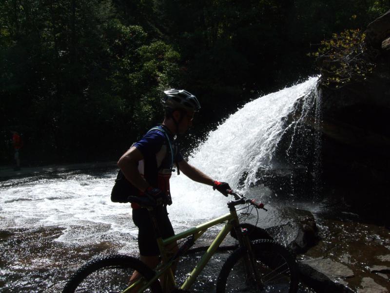 A silhouette of a mountain biker standing next to a waterfall, with water cascading over rocks in a lush green forest setting. The biker is holding a bicycle and wearing a helmet. Sunlight filters through the trees, creating a serene outdoor atmosphere. DuPont State Forest mountain bike trail.