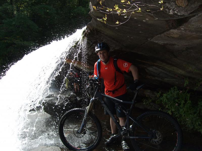 A mountain biker in an orange shirt and helmet stands next to a bicycle under a waterfall, with water cascading down rocks in a lush green forest setting. DuPont State Forest mountain bike trail.