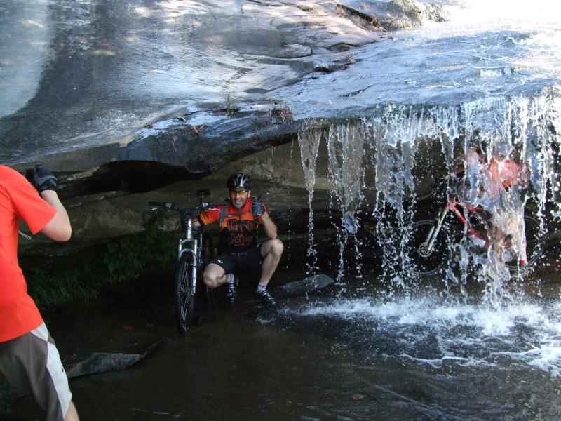 Two mountain bikers are enjoying a moment under a rocky overhang, sheltered from a waterfall. One biker, wearing a helmet and colorful jersey, is sitting on a rock with a bike nearby, giving a thumbs up. The second biker is partially obscured by the cascading water as they navigate their bike beneath the overhang. Sunlight glimmers on the water, creating a lively outdoor scene. DuPont State Recreational Forest mountain bike trail.
