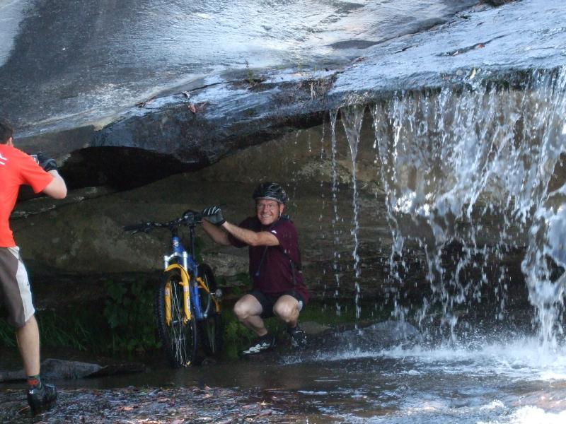 A man in a maroon shirt and helmet smiles while crouching under a rock overhang beside a mountain bicycle, with water cascading down in front of him. Another person in a red shirt is partially visible, interacting with the environment. The scene captures a moment of outdoor adventure near a waterfall. DuPont State Forest mountain bike trail.