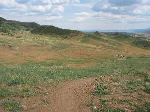 A landscape view showing a grassy plain with rolling hills under a partly cloudy sky. The foreground features a dirt path leading into the distance, bordered by patches of greenery and rocks, while the background showcases a serene, open terrain. Green Mountain mountain bike trail.