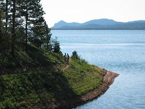 A scenic view of a tranquil lake surrounded by lush greenery and distant mountains. Two people are seen walking along the shoreline, enjoying the natural landscape on a sunny day. Rampart Reservoir mountain bike trail.