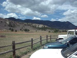 A scenic view of a grassy field framed by a wooden fence, with parked cars in the foreground and distant mountains under a partly cloudy sky. Ute Valley Park mountain bike trail.