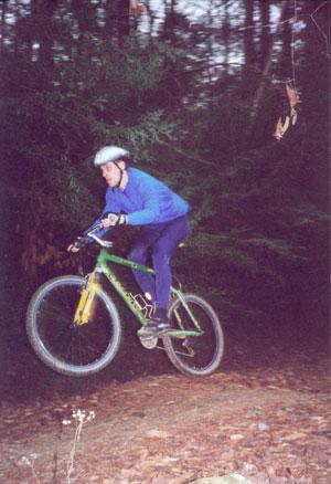 A person wearing a helmet and blue clothing is riding a green mountain bike while performing a jump on a dirt trail surrounded by trees. The scene captures the dynamic motion of mountain biking in a forested setting. Thrift Cove mountain bike trail.