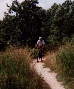 A cyclist riding along a dirt trail surrounded by tall grass and trees on a sunny day. Lake Crabtree County Park mountain bike trail.