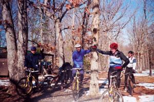 Four mountain bikers wearing helmets and cycling gear are gathered in a wooded area. They are standing beside their bicycles, surrounded by bare trees and a bit of snow on the ground. The sky is clear and blue. The group appears to be interacting, possibly discussing their ride. Country Park mountain bike trail.