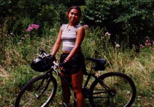 A smiling young woman standing next to a bicycle in a grassy area with wildflowers and trees in the background. She is wearing a sleeveless top and shorts, and she is holding onto the bike handles. Canada Creek mountain bike trail.