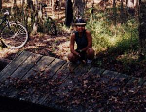 A person wearing a helmet squats on a wooden structure in a forested area, surrounded by trees and fallen leaves. A bicycle is visible in the background. Modoc (Stevens Creek Trail) mountain bike trail.