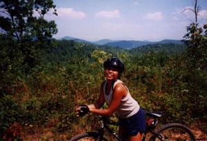 A person wearing a helmet leans on a mountain bike, smiling against a backdrop of green hills and a blue sky with scattered clouds. Stonewall Falls Loop mountain bike trail.
