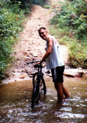 A young man standing in shallow water beside his mountain bike on a dirt trail surrounded by greenery. He is smiling at the camera, wearing a sleeveless white shirt and black shorts. The background features a rocky incline leading up the trail. Stonewall Falls Loop mountain bike trail.