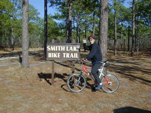A cyclist wearing a black jacket and red helmet waves while standing next to a sign that reads "Smith Lake Bike Trail," surrounded by tall pine trees and a gravel path. Smith Lake mountain bike trail.