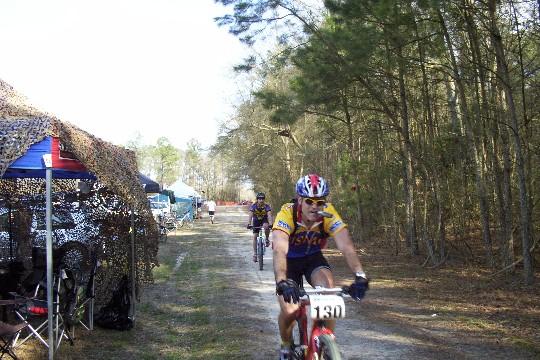 A mountain biker wearing a yellow and purple jersey and a helmet rides along a dirt path surrounded by trees. In the background, other cyclists can be seen, and there are tents set up on the side of the trail, indicating a biking event. The atmosphere is lively and outdoor-oriented. Bicycle Post Trails mountain bike trail.