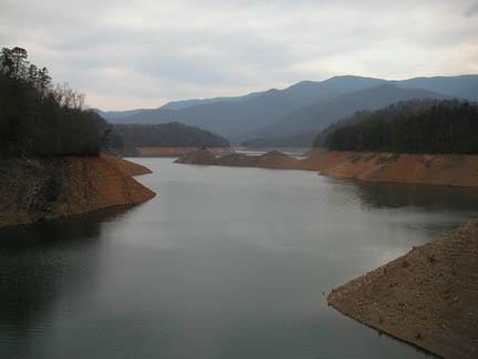 A scenic view of a tranquil lake surrounded by hilly terrain, with sandy banks and a backdrop of mountains under a cloudy sky. The water reflects the muted colors of the landscape, creating a serene atmosphere. Tsali Left Loop mountain bike trail.