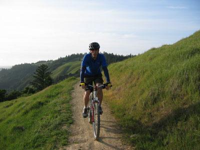 A cyclist wearing a blue jersey and a black helmet rides on a dirt trail surrounded by lush green hills and a clear sky. Saratoga Gap mountain bike trail.