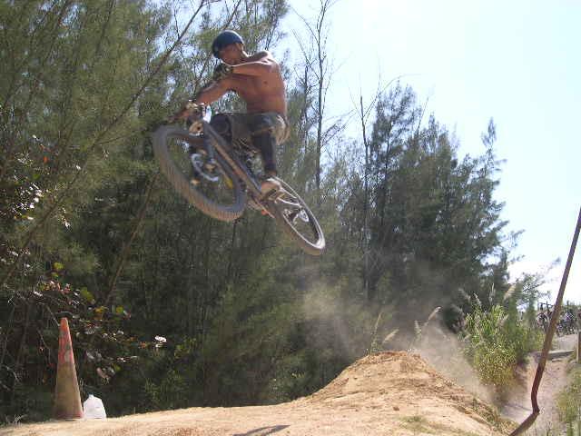 A shirtless cyclist performs a jump in mid-air on a mountain bike, surrounded by trees. Dust is kicked up from the dirt ramp as the rider lifts off, showcasing a dynamic moment of extreme sports. Oleta River State Park mountain bike trail.