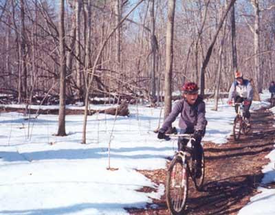 Two children riding bicycles on a muddy trail through a winter landscape, with patches of snow on the ground and bare trees surrounding them. One child is wearing a helmet and a jacket, focused on riding, while the other trails behind. The scene captures an outdoor adventure in a forested area. Country Park mountain bike trail.