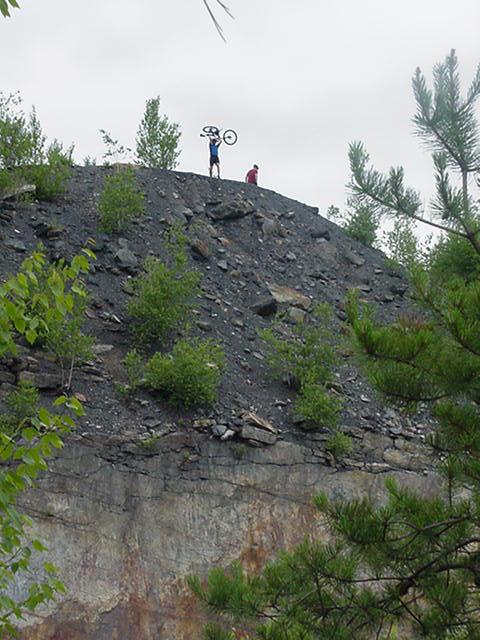 Two people are standing on a rocky hillside. One person is holding a bicycle above their head, while the other person is standing nearby, looking toward them. The scene is surrounded by greenery, with trees and shrubs partially covering the rocky terrain. The sky is overcast. Coal Cracker mountain bike trail.