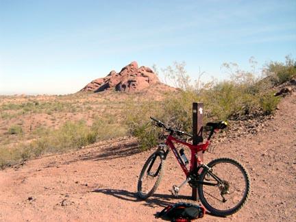 A mountain bike resting on a dirt trail in a desert landscape, with a prominent rock formation in the background and sparse vegetation surrounding the area. Papago Park Area mountain bike trail.