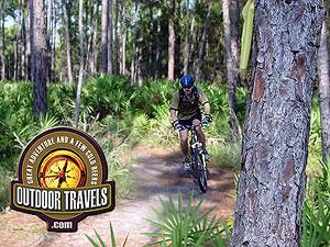 A mountain biker riding along a dirt path surrounded by lush greenery and tall trees, with the logo of "Outdoor Travels" at the bottom left corner. Morris Bridge Park mountain bike trail.