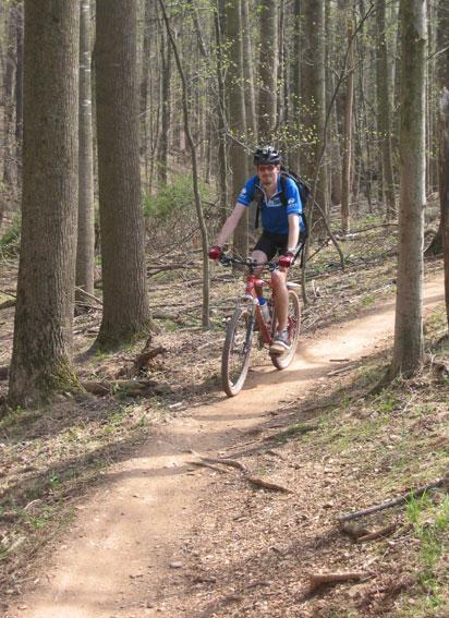 A person riding a mountain bike along a dirt path in a forested area. The cyclist is wearing a blue shirt and a helmet, surrounded by tall trees and greenery. The path is winding, indicating a natural trail through the woods. Schaeffer Farms mountain bike trail.
