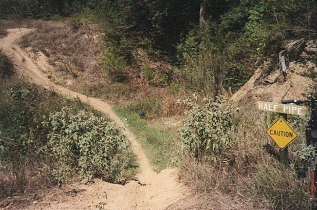 A dirt path leading to a half pipe structure, surrounded by overgrown vegetation and trees. A caution sign is visible next to the path. The area appears rugged and natural, indicating potential use for biking or skateboarding activities. Lake Bryan mountain bike trail.