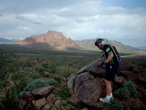 A person wearing a helmet and athletic clothing stands on a rocky outcrop, looking down at the terrain below. In the background, a mountain and expansive green landscape are visible under a partly cloudy sky. Hawes Loop mountain bike trail.