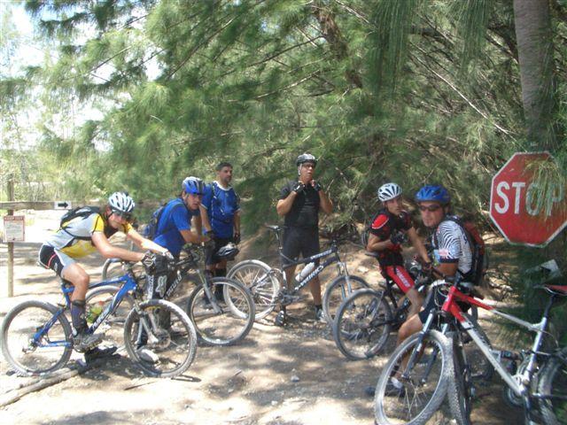 A group of cyclists, wearing helmets and various cycling attire, gather near a stop sign in a wooded area. They are on mountain bikes, some are seated while others are standing, engaged in conversation or preparing to continue their ride. Lush green trees surround them, indicating a natural outdoor setting. Oleta River State Park mountain bike trail.
