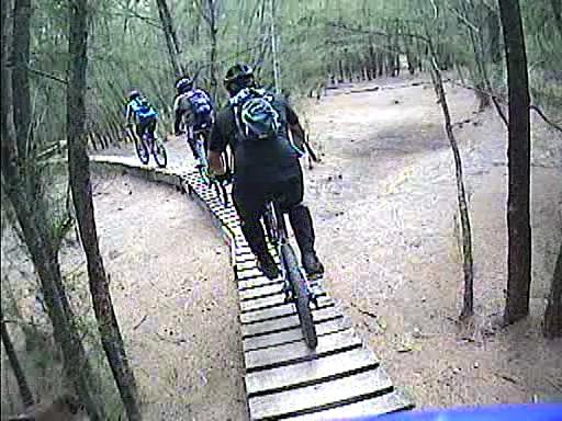 Three mountain bikers riding on a narrow wooden bridge through a forested area. The path is surrounded by trees, and the terrain is sandy. The bikers are dressed in activewear and wearing helmets, with backpacks visible. Oleta River State Park mountain bike trail.