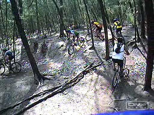 A group of mountain bikers navigating a dirt trail in a forested area, surrounded by trees. The riders are seen in various positions on the track, showcasing a mix of brightly colored clothing and bike gear. The trail is rugged, with visible roots and uneven ground. Oleta River State Park mountain bike trail.