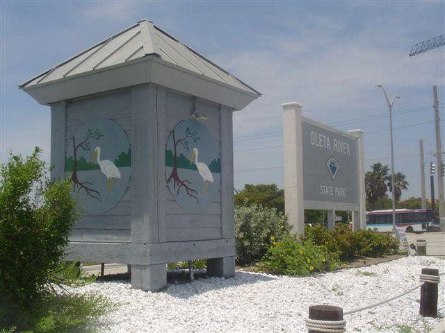 Signage for Oleta River State Park, featuring a decorative structure with bird paintings and a large sign indicating the park's name, surrounded by greenery and white gravel landscaping. Oleta River State Park mountain bike trail.
