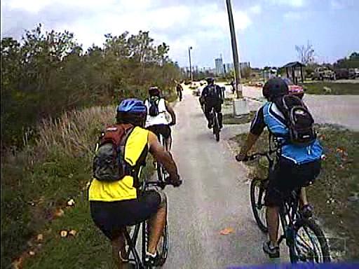 A group of cyclists riding on a paved path surrounded by greenery. The cyclists are wearing helmets and backpacks, and the scene includes a mix of bicycles and distant buildings. The weather appears clear with a few clouds in the sky. Oleta River State Park mountain bike trail.