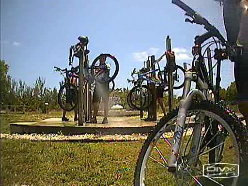 A bike rack in a park with several bicycles parked. One person is adjusting a bike near the rack under a clear blue sky. Green vegetation can be seen in the background. Oleta River State Park mountain bike trail.