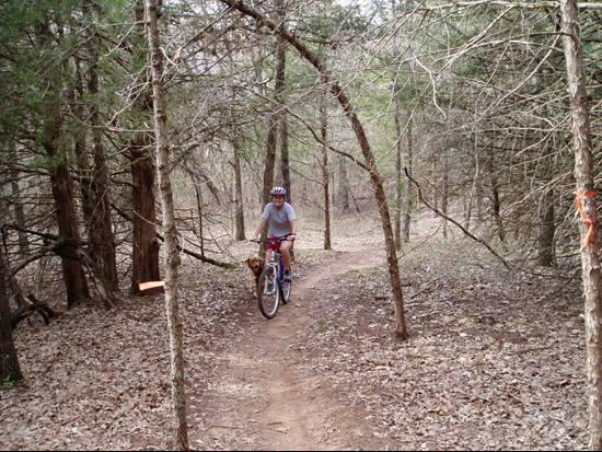 A person riding a bicycle on a dirt trail surrounded by trees in a forested area. The path is narrow with visible leaf litter, and there is a small signpost with an orange marker on the left. The scene conveys a sense of outdoor adventure and nature exploration. McMurtry Trail mountain bike trail.