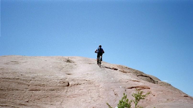 A mountain biker ascends a rocky terrain under a clear blue sky. The biker is wearing a helmet and riding uphill on a smooth, light-colored surface, with green shrubbery visible in the foreground. Slickrock mountain bike trail.