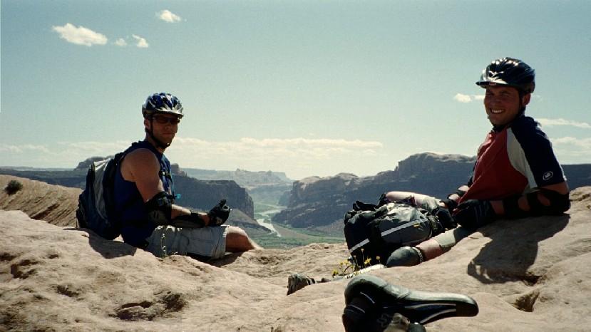 Two mountain bikers sitting on a rocky ledge, overlooking a scenic valley. One is wearing a blue sleeveless shirt and a helmet, while the other is in a red and white shirt and a helmet. The background features a river meandering through the landscape, with cliffs and blue sky above. Slickrock mountain bike trail.