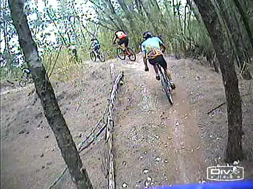 A group of mountain bikers riding on a dirt trail surrounded by trees, with one cyclist in the foreground performing a jump. The scene captures the excitement and action of the sport in a natural outdoor setting. Oleta River State Park mountain bike trail.
