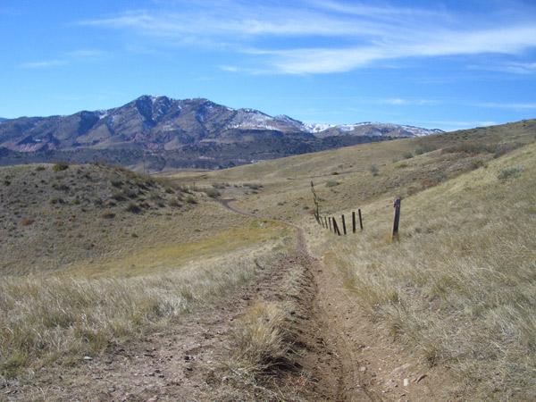 A winding dirt path cuts through grassy hills, leading toward a mountainous landscape in the distance. Snow-capped peaks are visible under a clear blue sky, with scattered clouds. Wooden fence posts line the right side of the trail, enhancing the natural scenery. Green Mountain mountain bike trail.
