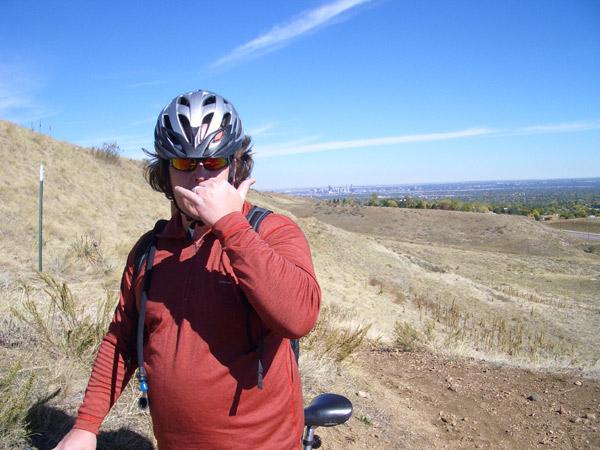 A person wearing a bicycle helmet and sunglasses stands on a grassy hillside, gesturing with one hand while covering their mouth with the other. The background features a view of distant city buildings against a clear blue sky. Green Mountain mountain bike trail.