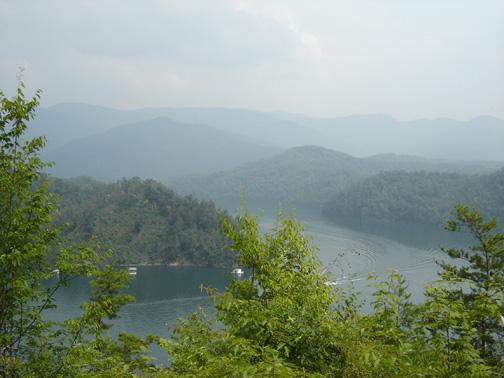A panoramic view of a serene lake surrounded by lush green hills and mountains under a hazy sky. The water reflects the nearby trees and distant peaks, creating a tranquil natural landscape. Tsali Mouse Branch Loop mountain bike trail.