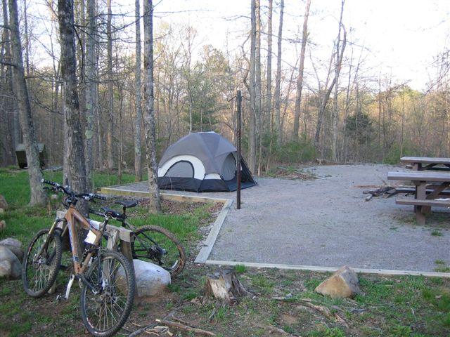 A campsite scene featuring a black and gray tent set up in a wooded area. Two mountain bikes are parked nearby on gravel, with a picnic table visible in the background. The setting is surrounded by trees and greenery, indicating an early spring or late afternoon atmosphere. Chilhowee trail system mountain bike trail.