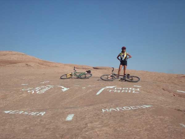 A mountain biker standing on rocky terrain with two bicycles beside them. The ground features painted trail markers indicating "Easier" and "Harder" trails, with a clear blue sky in the background. Slickrock mountain bike trail.