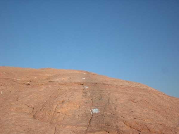 A rocky slope under a clear blue sky, showing distinct layers of sedimentary rock with a few white markings visible on the surface. Slickrock mountain bike trail.