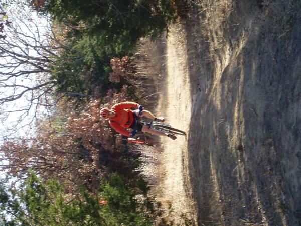 A cyclist in an orange shirt is riding a mountain bike on a dirt trail surrounded by trees with autumn foliage. The scene captures the cyclist in motion, facing forward, with a slightly elevated position above the ground. The background features a mix of green and brown foliage, indicating a natural outdoor setting. McMurtry Trail mountain bike trail.