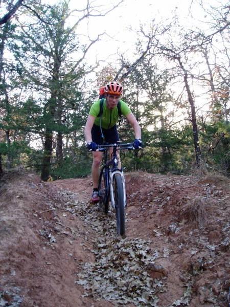 A mountain biker navigating a rocky dirt trail surrounded by trees, wearing a bright orange helmet and a green shirt. The cyclist appears focused as they ride over a path covered with fallen leaves, with the sun setting in the background. McMurtry Trail mountain bike trail.
