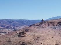 A lone mountain biker rides along a rocky trail with expansive mountainous landscapes in the background under a clear blue sky. Slickrock mountain bike trail.