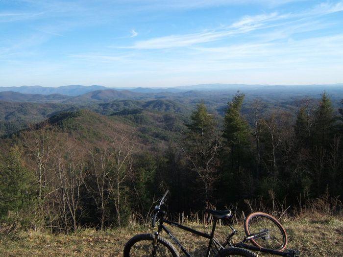 A scenic view of rolling mountains under a blue sky, with two bicycles resting on a grassy area in the foreground. The landscape features a mixture of evergreen and deciduous trees, stretching across the horizon. Bear Creek mountain bike trail.