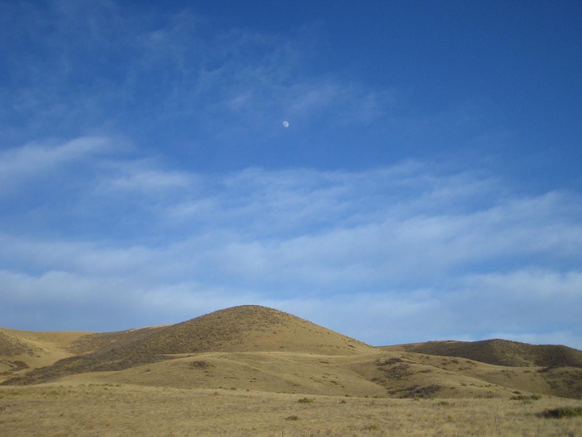A serene landscape featuring rolling hills under a clear blue sky, with wispy clouds and a visible moon in the background. The dry, grassy terrain showcases muted earth tones. Green Mountain mountain bike trail.