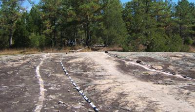 A rocky terrain with visible natural pathways, surrounded by dense green trees and vegetation under a clear blue sky. The landscape features smooth, worn surfaces indicative of erosion and time. Georgia International Horse Park mountain bike trail.
